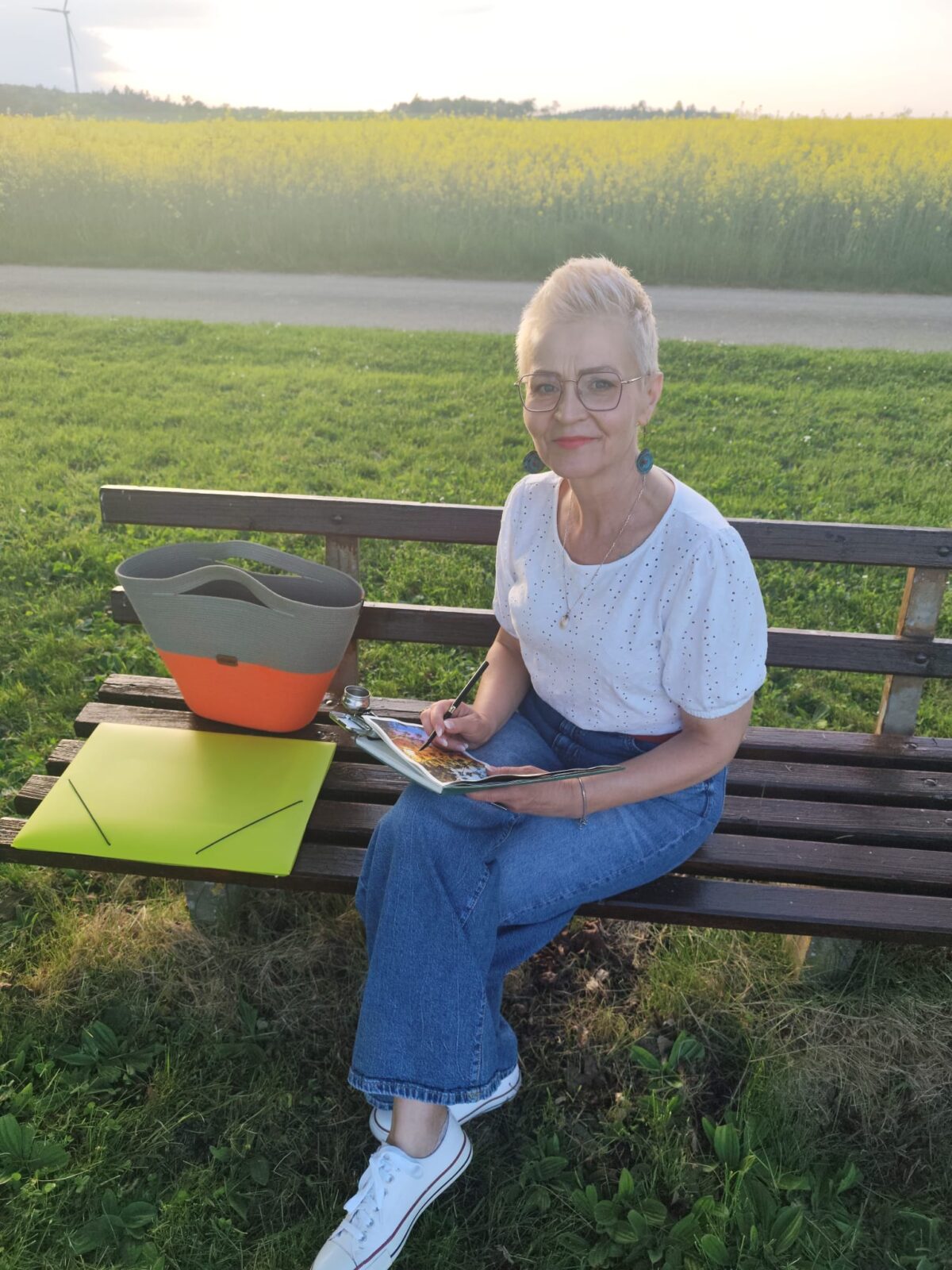 Monika Bargieł beim Malen im Freien, sitzend auf einer Bank mit Skizzenbuch und Aquarellfarben.